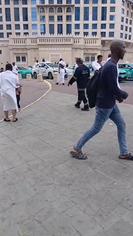 The video depicts a paved area outside a tall building, possibly a hotel, where several people are walking around. Most of the men are wearing white robes. A man in a white robe is holding the hand of a young girl in a pink dress. A person in a dark uniform with reflective striping is walking in the opposite direction of the man and girl. There are cars parked nearby, including green sports cars and white taxis. The building behind the people has many windows and a decorative top.