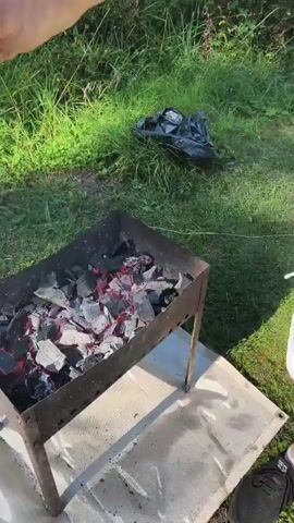 In an outdoor setting with grass and foliage, a person wearing a white shirt and shorts is standing to the right of a metal barbecue grill. The grill contains glowing red coals. The person is holding a black shovel in their right hand and appears to be pouring something from a clear plastic container into the grill. To the left of the grill, there is a black plastic bag on the ground. The person's left hand is visible and holding the plastic container. The video focuses on the action of pouring into the grill, with the person's face not visible. The ground around the grill is covered by a light-colored tarp.