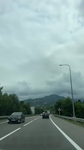 A first-person perspective video shows a car driving on a highway under a cloudy sky. Lush green trees line both sides of the road, and in the distance, rolling hills covered in vegetation are visible. Several other vehicles, including cars and a truck, are also on the highway. The road has clear white lane markings. A traffic sign with a warning symbol of a falling rock is positioned on the right side of the road. The video captures the drive towards a tunnel in the distance.