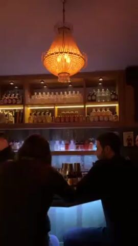 A dimly lit bar with a chandelier above features two people seated at the bar. A woman with reddish hair sits to the left, and a man with dark hair sits to her right. Behind the bar is a shelving unit filled with various bottles, illuminated by warm light. The bar itself is lit with blue light. A man is partially visible to the far right. The camera angle focuses on the two people at the bar from behind.