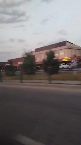 The video shows a street scene with a commercial building on the right side of the road. The building has multiple storefronts and a large sign that reads "AVECKET". There are trees and cars in front of the building, and the sky is cloudy. The video appears to be taken from a moving vehicle, as indicated by the slight blur and the perspective. The lighting suggests it is either early morning or late evening.
