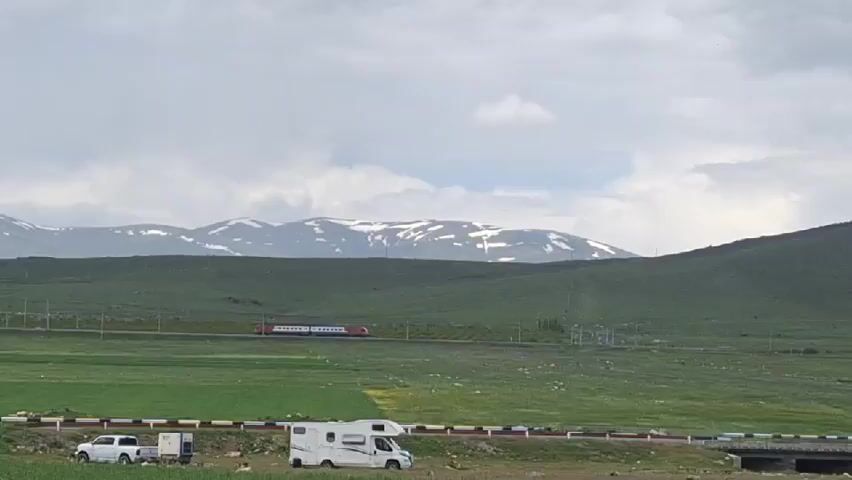 The scene is an outdoor shot of a green field with mountains in the background. The mountains have patches of snow on them. A train is traveling horizontally across the field.  In the foreground, there is a white truck with a trailer and a white RV parked next to a road. The sky is cloudy. A burgundy car is driving past the truck. The train is the most prominent object in the scene.