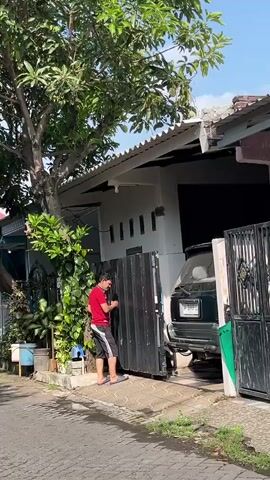 A man wearing a red t-shirt and striped shorts is closing a black gate in front of a house. The gate is partially open, and the man is pushing it closed. There is a dark green car parked in the driveway behind the gate. A tree with green leaves is to the left of the gate. The ground is wet, and there are some plants in pots near the gate. The sky is blue with some clouds. The scene appears to be outdoors, possibly in a residential area.