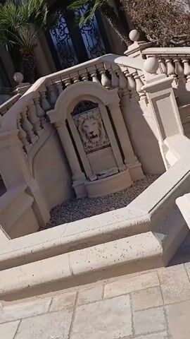The video shows the exterior of a large, opulent house on a sunny day. The house is white with beige trim and features arched windows with dark frames and wrought iron details. Two small palm trees flank the entrance. A decorative stone staircase with a lion's head fountain leads up to the entrance. The stairs are made of light-colored stone with ornate balusters. Vines climb up the walls of the house. The ground is paved with square stones.