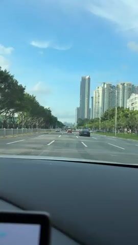 A view from inside a car driving on a road. There are trees lining the road on both sides. In the background are tall buildings. The car's dashboard is visible with a screen showing a GPS navigation map. The map indicates the car is traveling at 59 km/h and will arrive in 8 minutes. There is also a camera mounted on the windshield. The steering wheel is white and black.