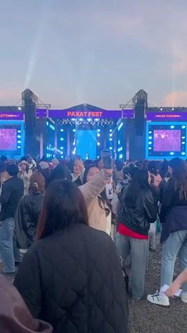 The video shows a large crowd of people attending an outdoor music festival called Paxat Fest. The main stage is visible, featuring large screens displaying the performing artist, RaiM. The stage is decorated with a lighting system and the festival name in large letters. The crowd is diverse, with people of different ages and ethnicities. Many are holding up their phones to record the performance, while some are dancing and enjoying the music. The sky is slightly overcast, suggesting it might be evening. The general atmosphere appears lively and festive.