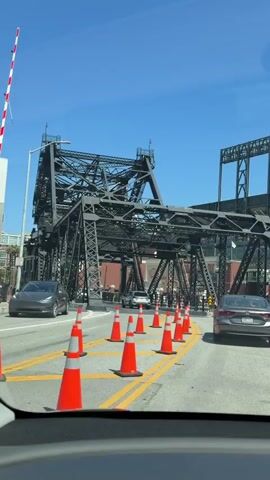The video shows a black steel truss bridge from the perspective of someone in a car. The car is driving on a road with orange traffic cones and yellow lane markings. Several cars are also on the road. The sky is blue and clear, and buildings can be seen in the background. The bridge appears to be a drawbridge or a similar movable structure, as there are visible mechanisms for lifting or opening it. The overall scene suggests that the area may be undergoing some construction or has temporary traffic changes in place.
