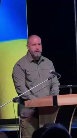 The video shows a bald man with a gray beard speaking from behind a wooden lectern. He wears a light green collared shirt. The backdrop is a large screen showing the Ukrainian flag. Two microphones are positioned on the lectern. The man looks directly at the camera and gesticulates with his hands as he speaks. The lower portion of the frame reveals silhouettes of people listening to the speech.