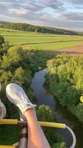 The video shows a view from a hot air balloon basket. The basket railing is visible in the foreground, with a person's leg and white sneaker resting on it. Below, a winding river flows through a lush green landscape. The riverbanks are densely covered with trees and vegetation. Beyond the river, there are large, flat green fields, possibly farmland, stretching towards a tree-lined horizon. The sky is blue with scattered white clouds, and the lighting suggests it is late afternoon or early evening.