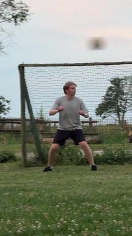 This media file features a young person in athletic wear standing in front of a soccer goal. They are wearing a light grey t-shirt and black shorts with white accents on their shoes. Their stance suggests they are anticipating or actively participating in a soccer-related activity. The background shows a grassy field with scattered white wildflowers, a wooden fence, and trees. The sky appears overcast, indicating a cloudy day. The overall setting appears to be an outdoor recreational area, possibly a park or a field near a rural or semi-rural location. The lighting suggests it is likely late afternoon or early evening. No visible text is present in the media file. The scene conveys a sense of casual sport and outdoor activity.