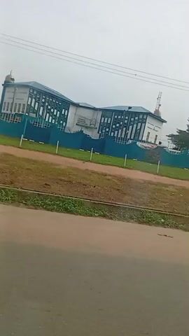 A moving vehicle passes a building. The building has a white facade with blue and white details. It is surrounded by a green fence and a lawn. A tree is visible behind the fence. The sky is overcast. The road is paved.