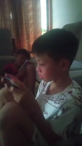Two young boys are sitting on the floor in a dimly lit room, each holding a smartphone and looking at the screen. The boy in the foreground, wearing a white t-shirt with a maroon design, is closer to the camera and is shown in profile. He is holding the phone with both hands and appears to be actively engaged with it. The second boy, in the background and slightly out of focus, is wearing a red shirt and is also looking at his phone. He is sitting with his legs crossed. In the background, there is a window with sheer curtains and a couch. The overall impression is one of quiet activity, with both children absorbed in their electronic devices.