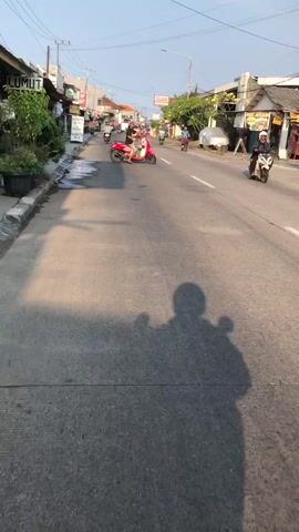 The media shows a street scene in Boro, East Java, likely in the morning or late afternoon due to the long shadows cast by the setting or rising sun. The weather appears to be clear and sunny.

The primary subjects are people riding motorcycles. In the foreground, a person is riding a black motorcycle, seen from behind. They are wearing a helmet, a dark jacket, and a backpack with a design on the back. Their shadow is prominent on the road. In the middle ground, a person on a red scooter is also visible, wearing a light-colored top and a helmet. Other motorcycles and riders are present further down the street.

The setting is a paved road with buildings lining one side. These appear to be shops or small businesses. Some of these buildings have signs. One sign, partially visible on the right side, has text that includes "AYAM". Another sign on the left has text like "JALAN". There's also a sign with a blue background and white text, partially obscured. The road has markings and some patches of wetness or oil stains. The overall atmosphere seems like everyday life in a moderately busy area.