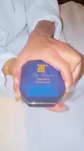 A person wearing a white robe opens a small, blue Joy Treasurers Jewellers jewelry box to reveal a pair of square earrings with dark blue gems and white settings. The person is holding the box while sitting on a white bed.