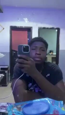 A young, dark-skinned man is taking a selfie in a mirror. He is wearing a black t-shirt with white writing on it and has short, dark hair. He is holding a dark-colored phone in his right hand, and a white charging cord is plugged into the phone. The room is lit with a blueish light. Behind him in the mirror, there are two doorways. The doorway on the left has a red towel hanging behind it. A package of blue wipes is visible on the surface in front of the mirror.