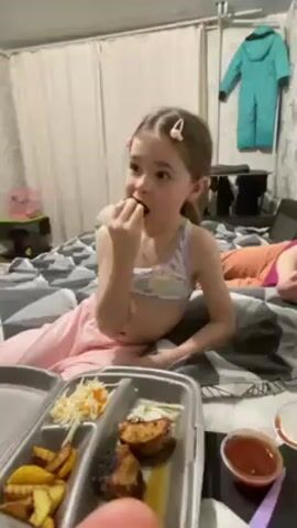 A young girl with light brown hair, parted to the side and held with pink clips, is seated on a bed with a grey and white patterned comforter. She is wearing a light-colored top with a pattern and pink pants. She holds a piece of meat in her hand, which she is eating. In front of her is a grey divided food container with fries, shredded cabbage, and two pieces of meat. There is a small container of red sauce next to the food. Behind her, another person is lying on the bed, partly visible in an orange top. The room has a light-colored curtain, wallpaper, and a turquoise snowsuit hanging on the wall.