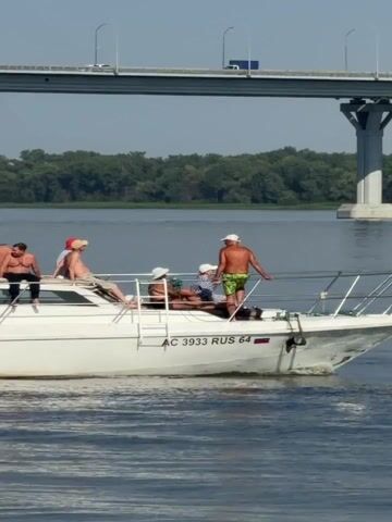 A white motorboat with a cabin is cruising on a body of water. Six individuals, appearing to be adults, are on board. Three men are seated on the port side edge of the cabin's roof, facing towards the left. One man is leaning against the cabin, another is sitting with legs extended, and the third is leaning forward. On the starboard side, towards the stern, another man stands near the railing, facing the left. Two other individuals are lying down on the deck of the boat, one with a hat on. The boat has visible registration numbers on its side: "+7 927 223 92 23" and "AC 3933 RUS 64". In the background, a large concrete bridge spans across the water. Trees and foliage line the distant shore. The sky is clear and blue. The water is rippled, indicating the boat is in motion.