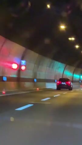 The video shows a view from inside a car driving through a tunnel at night. The tunnel has a curved ceiling with lights running along it. The walls of the tunnel are illuminated with alternating red and green lights. There are blue square lights embedded in the walls at regular intervals, and red circular markers on the road surface. A black SUV is driving ahead in the same lane. The road is marked with white dashed lines. There are also some digital signs visible, one showing "112-HVALA. POZIV 112. EMERGENCY CALL NUMBER". Another car, an orange sedan, is visible in the right lane. The video is slightly blurry and has some bokeh effects from the lights.