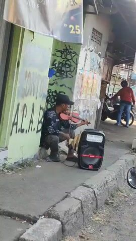 A person is sitting on the sidewalk playing a violin. They are wearing a black baseball cap, a patterned shirt, and dark pants. Next to them is a black speaker with a red accent. The person is leaning against a building that has graffiti on it, including the letters "A.C.A.B.". There is a curb and a street in front of the sidewalk. In the background, another person is standing next to a red motorcycle. The scene appears to be outdoors during the daytime.