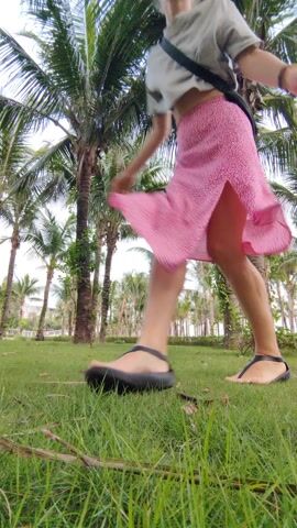 An adult woman with light-colored hair, styled in an updo, is captured in a two-frame sequence showcasing joy and movement in a tropical setting. She wears a light gray or off-white short-sleeved top, a flowing pink midi-skirt with a subtle vertical texture, and black flat sandals. A dark (likely black or deep green) fanny pack or small crossbody bag is worn across her chest. A bracelet is visible on her left wrist.

In the first frame, she walks away from the camera, looking back over her left shoulder with a smile. In the second frame, she is mid-jump or mid-spin, with her arms outstretched and a wide, happy smile directed towards the camera.

The scene is a lush green grassy area, viewed from a low angle, placing the viewer almost at ground level. The background features a dense cluster of tall palm trees with slender trunks and full green fronds, suggesting a well-maintained park, garden, or resort. Faintly visible light-colored buildings or structures can be seen in the distant background beyond the trees. The sky is bright and appears overcast or hazy white, indicating daytime. Some dried brown plant debris, such as twigs and leaves, is scattered on the grass in the immediate foreground. A white rectangular label with unreadable black text is affixed to the trunk of the palm tree on the far right. The overall impression is one of happiness and freedom, consistent with a leisurely outdoor activity in Bai Chay Ward, Vietnam.