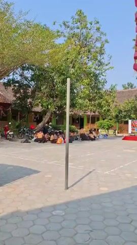 A medium shot captures an outdoor courtyard in daylight, with a tall, thin metal pole standing vertically in the foreground, casting a shadow on the hexagonal paved ground. In the middle ground, a group of children wearing brown uniforms, possibly scouts, are sitting on the ground in a semicircle under the shade of a large tree. Some of the children are looking towards the left, while others are interacting with each other. Bags and a red motorcycle are visible near the children. Behind the children, a building with red-tiled roofs and green accents can be seen. The overall environment appears to be a school or community gathering space on a sunny day.