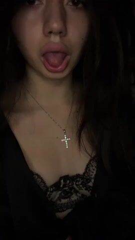 This is a close-up, vertical video of a person's face and upper body. The person has long dark hair and is wearing a black top with lace detailing and a silver necklace with a jeweled cross pendant. Their tongue is sticking out. The background is dark and indistinct.