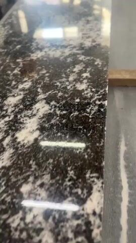 This video shows a close-up, moving shot of a granite countertop. The granite has a speckled pattern of black, white, and grey. The countertop is polished and reflects light. The camera is moving slowly across the surface of the countertop, creating a slightly blurred effect. In the background, there appears to be a wooden object and possibly some industrial equipment. The lighting in the scene is bright, with reflections visible on the countertop. The video seems to be shot in a workshop or showroom environment.
