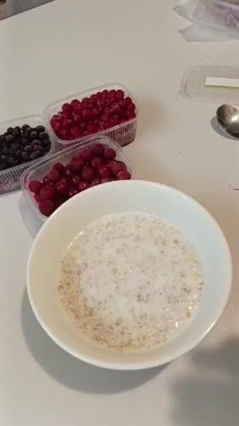 A person is adding red berries to a bowl of oatmeal. There are three containers of berries on the table. One container has black berries, another has red berries, and a third, also with red berries, is being held by the person. The oatmeal is in a white bowl and appears to have milk in it. The background is a white surface, likely a table.
