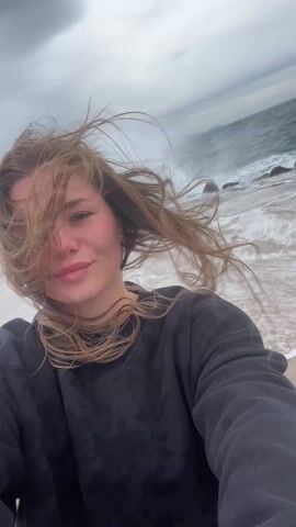 A young woman is filming a selfie video on a beach. She has long blonde hair being blown by the wind and is wearing a black sweatshirt. The ocean is behind her with waves crashing on the shore, and mountains are visible in the distance under a cloudy sky. The camera is held at arm's length, and the woman is smiling and looking at the camera.