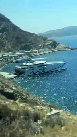 A white and blue high-speed ferry named SEA JETS is docked at a port. The ferry is large and has multiple decks. The port is located on a rocky, mountainous coastline with a clear blue sky above. The water is a vibrant blue and appears somewhat choppy. Other boats and structures are visible in the background at the port. The camera perspective is from a higher vantage point, looking down at the scene.