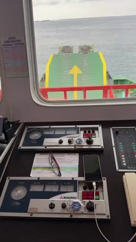 The video shows the interior of a ship's bridge, looking out a large window at the ocean and the ship's deck. The bridge is equipped with various control panels, gauges, a steering wheel, and a compass. In the foreground, there are two large control consoles with levers, buttons, and screens. A pair of glasses and a smartphone are resting on one of the consoles. A schedule titled "Jadwal Garda Maritim" is attached to the wall. Further back, a man wearing a captain's hat and uniform is visible, facing away from the camera and looking at a monitor. The view outside the window reveals a calm sea under an overcast sky, with the rear of the ship's deck visible. The overall impression is one of a working vessel in operation.