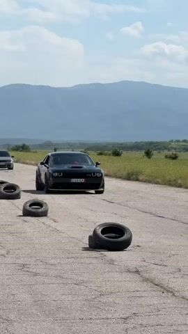 The video shows a black Dodge Challenger stopped on an asphalt road with a mountain range in the background. There are several tires lying on the road.  Two men are walking to the right of the car. The car has a license plate that reads "016 B 424" and a logo that reads "BIDMOTORS". The sky is blue with some clouds, and the surrounding area is fields of green grass.