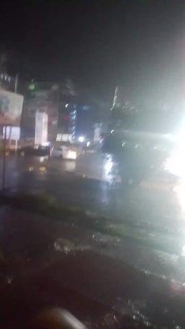 The video depicts a rainy night scene in an urban area. Streetlights and neon signs illuminate the wet pavement and a few parked cars. A small tree is visible in the foreground. The camera is unsteady, and the image is slightly blurry.