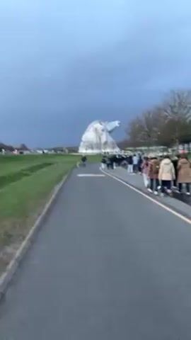 The video shows a paved path leading towards The Kelpies, two large horse-head sculptures in Scotland. There are a number of people walking along the path towards the sculptures, as well as a person walking a dog. On the left of the path is a grassy bank. In the background, power lines can be seen. The sky is overcast.