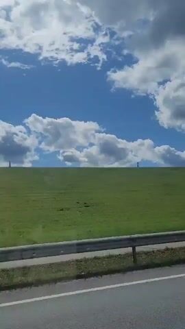 The video shows a grassy hillside under a blue sky with white clouds. There are two poles visible on top of the hill. A metal guardrail runs along the bottom of the frame, separating the hill from a road. The camera appears to be moving, as evidenced by the slight blurring and shift in perspective.