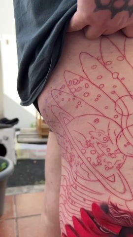 A person wearing a dark gray T-shirt stands on a brick patio, holding up the fabric to reveal a tattoo on their left thigh. The tattoo features a red and black abstract image of a face in the lower half, with red line art of a planet and organic shapes above. A few objects such as a plant pot, a white box, a drying rack, and a partial view of a bench are visible in the background. The person remains stationary, allowing a clear view of the tattoo design.
