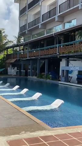 A wide-angle shot shows a swimming pool with built-in white lounge chairs submerged in the water. The pool is in the foreground, with a hotel building in the background. The hotel has multiple levels with balconies and rooms. There are palm trees visible to the left of the building. A "WELCOME" sign is visible on a white structure near the pool. The sky is cloudy. There are no people or animals visible in the video.