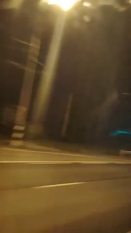 This is a video compilation of a nighttime drive on a road, likely a highway, in Zavidovo, Russia. The footage is shaky, indicating movement, possibly from a vehicle. The scene is dark, with streetlights and passing lights creating streaks of illumination across the road and surrounding trees.

The primary subjects are the road itself, characterized by yellow and white lane markings, and the dark, blurry silhouettes of trees and occasional structures along the roadside. A metal guardrail is visible separating the road from the adjacent area. The lighting suggests it is nighttime, and there are no clear indications of weather conditions, though the darkness and artificial lights are prominent.

The activity is clearly a drive. The fast-moving streaks of light and the blurred landscape convey a sense of speed and transit. There are no people or specific objects clearly identifiable in the frame beyond the road infrastructure and natural elements. The overall emotion conveyed is one of movement through darkness, with the artificial lights offering brief moments of visibility. No visible text is present in the video.