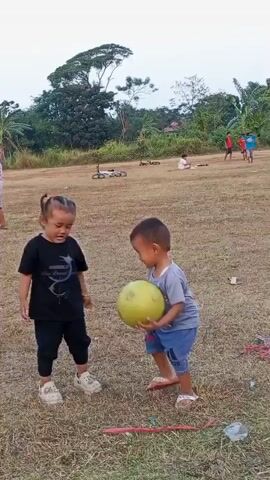 Two young children are playing in a grassy field. A child on the left, wearing a black t-shirt and black pants, is looking towards the right. A child on the right, wearing a grey t-shirt and blue shorts, is holding a yellow ball. In the background, there are several other children playing and some bicycles lying on the ground. The environment appears to be an outdoor field with trees and vegetation in the distance. The lighting suggests it is either early morning or late afternoon.