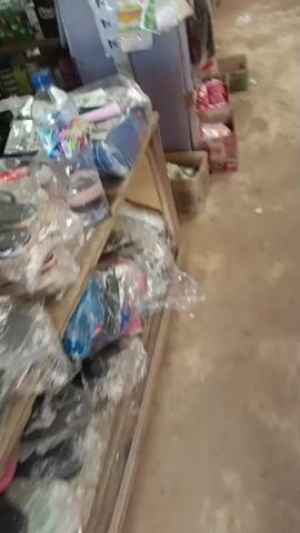 The video appears to be filmed from a moving camera, possibly held by a person walking down a narrow, dimly lit corridor. The floor is concrete and appears somewhat dirty and uneven. On the left side of the corridor, there are several cardboard boxes, some open and containing items that look like packaged goods. Some of these packages are brightly colored. Further down the corridor, on the right side, a white plastic stool is visible. Near the end of the corridor, leaning against the wall, are what appear to be cleaning tools, such as brooms or dustpans, some of which are wrapped in plastic. The overall impression is of a storage area or a back room. The video is shaky and the focus is not consistently sharp, suggesting it was filmed quickly and without stabilization.