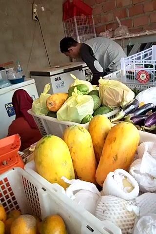 The video shows a market stall with a variety of fresh produce. In the foreground, there are baskets filled with papayas, oranges, and other fruits. Behind these, more baskets contain eggplants, chilies, and other vegetables. A person is visible in the background, seemingly arranging or packing produce. There is also a refrigerator and shelves stocked with more food items. The overall setting appears to be an outdoor or semi-open market.