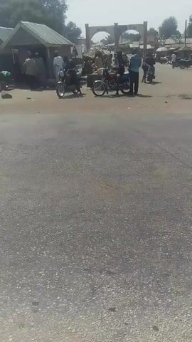 The video shows a rural street scene in what appears to be a West African country. The road is asphalt, and there are several motorcycles parked on both sides of the road. Several people are standing around talking, and one motorcycle is driving away. There are small shops or stalls along the side of the road, and a large archway stands in the background, bearing what seems to be the name of the city or town.  A blue car is also parked on the right side of the road. A billboard with a person's image is also seen on the right side of the road. The weather appears to be sunny.
