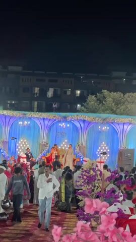 The scene is a nighttime outdoor event, possibly a wedding or a formal gathering, with a large stage decorated with blue curtains, chandeliers, and light fixtures. A couple is seated on the stage. The man is wearing a suit, and the woman is wearing a gown. The stage is surrounded by people, many of whom are walking around and looking at the stage. Some people are wearing traditional Indian clothing, suggesting the event may be culturally significant. There are floral decorations in the foreground. The background shows a residential building with multiple floors. The overall impression is a festive and celebratory atmosphere.