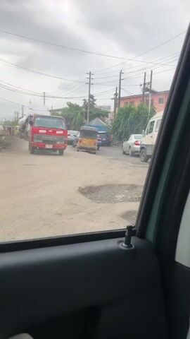The video begins from the interior of a vehicle, showing the view through the passenger side window. Outside, a red Toyota truck is visible on a dirt road, followed by a yellow three-wheeled auto-rickshaw, and then a white truck. The road appears to be in poor condition with several potholes. Utility poles and wires line the side of the road, with a yellow wall and some debris behind it. The sky is overcast. The camera briefly pans to the interior of the vehicle, showing the door panel and a white object covered in plastic. The video then returns to the exterior view, showing the same scene of the road with the vehicles. The white truck in the distance appears to be a large semi-trailer truck. The auto-rickshaw and the red truck are moving away from the camera. The video ends with the white truck in the distance, with no significant changes in the scene.