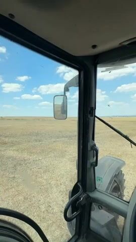 This video offers a POV perspective from inside a tractor cab, looking out at a dry, harvested field under a partly cloudy blue sky. The camera pans and shifts slightly, revealing more of the interior of the cab, including the steering wheel, control levers, and dashboard. A side mirror reflects the interior and a person looking at their phone. Outside the cab, the landscape appears flat with sparse vegetation and some hay bales visible in the distance. The video captures the agricultural setting and the experience of being inside a working tractor.