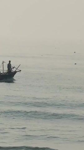 A fishing boat with two people on board motors across the water. The boat is small and dark, with some equipment visible on it. One person sits at the back of the boat, while the other stands at the front, possibly steering or observing. The water is gray and slightly choppy with small waves. The sky is overcast and hazy, creating a muted and indistinct background.