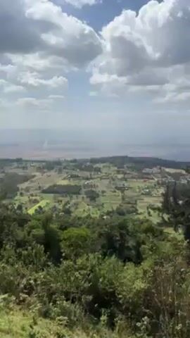 The video shows a panoramic view of a lush, green landscape under a partly cloudy sky. The camera is positioned high up, likely on a hillside or elevated viewpoint, overlooking a valley filled with trees, fields, and scattered buildings. The landscape stretches out towards distant hills or mountains on the horizon. The video appears to be taken during the day, with good natural lighting. The overall environment looks peaceful and rural. The voices of people speaking in the background can be heard, but they are not visible in the frame.