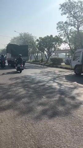 The video shows a roadside scene in daylight. In the foreground, the asphalt road is visible, marked with faint tire tracks and scattered debris. Tree shadows are cast across the road from the right. On the right side of the road, a large tanker truck is parked. To the left of the road, a scooter with a rider is traveling away from the camera. Behind the scooter, another truck with a green tarp covering its cargo is also moving away. Trees and foliage line the side of the road, with a utility pole visible in the distance on the left. The sky is clear with sunlight filtering through the trees.