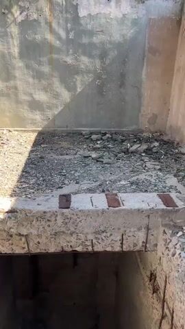 A handheld camera pans across the interior of a severely dilapidated building, likely abandoned and possibly undergoing demolition. The structure is made of concrete and brick, with extensive damage evident. Walls are cracked and peeling, exposing rebar and rubble. Floors are covered in debris, including loose concrete chunks, gravel, and rusted metal. In some areas, open pits or drop-offs are visible, indicating missing floors or stairwells. The scene is stark and industrial, with natural light filtering in through openings and casting shadows. There are no people or animals visible, and the focus remains on the crumbling architecture and the overall sense of decay and neglect.