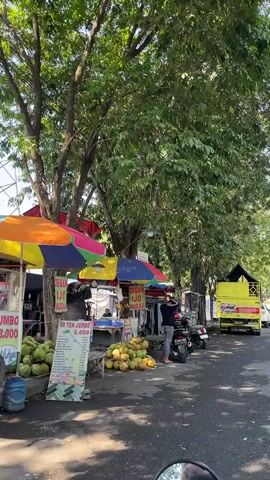 The video shows a roadside scene with several vendors and their stalls set up under trees. The stalls have colorful umbrellas and display various goods, including fruits like melons and drinks advertised on a sign. People are seen interacting with the vendors. There are also motorcycles parked and a yellow truck moving on the road in the background. The environment appears to be a sunny day, with dappled sunlight filtering through the leaves of the trees.